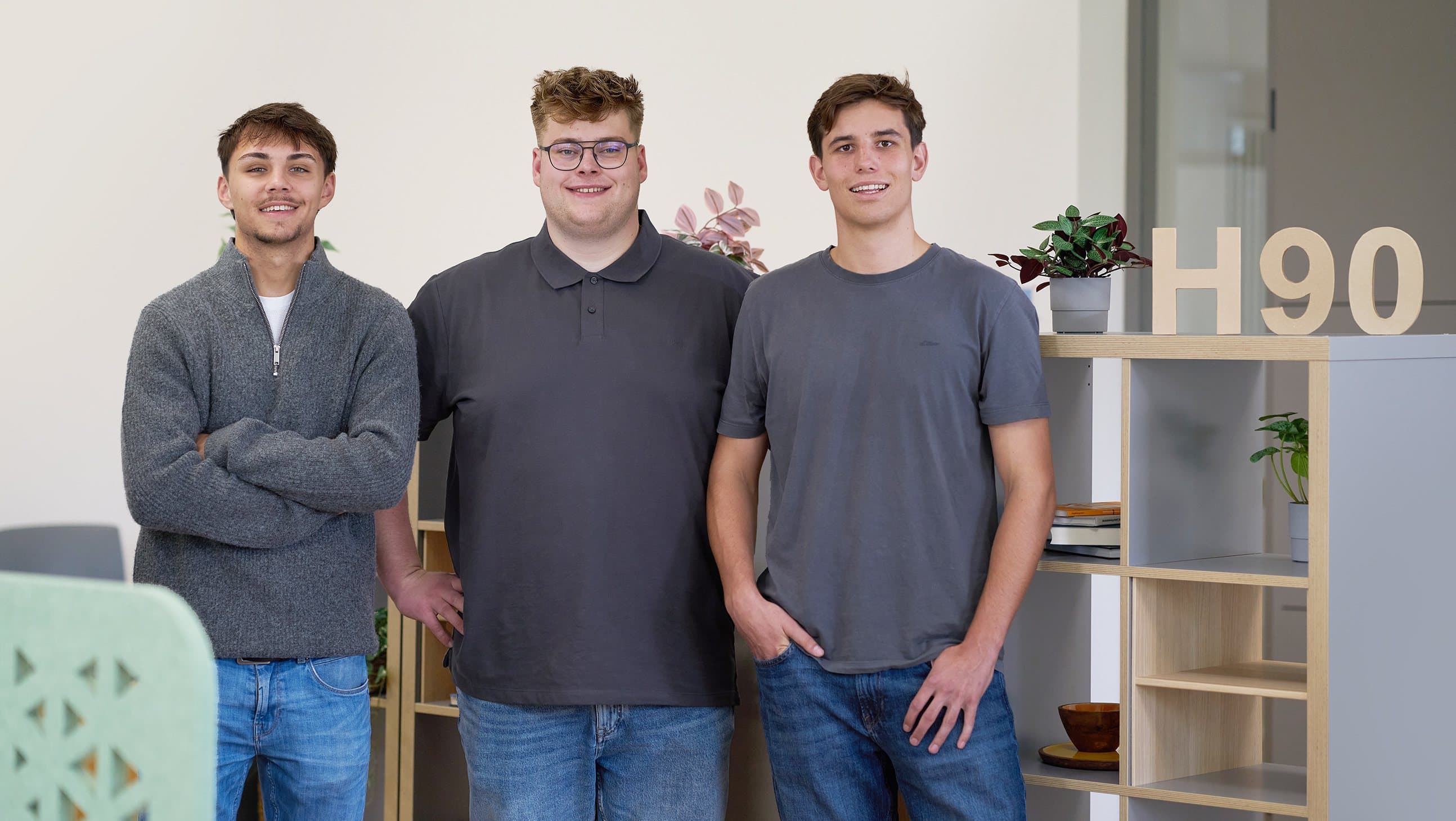 Luis Sabisch, Paul Budde und Max Wiebe stehen nebeneinander im Büro vor einem hellen Regal.