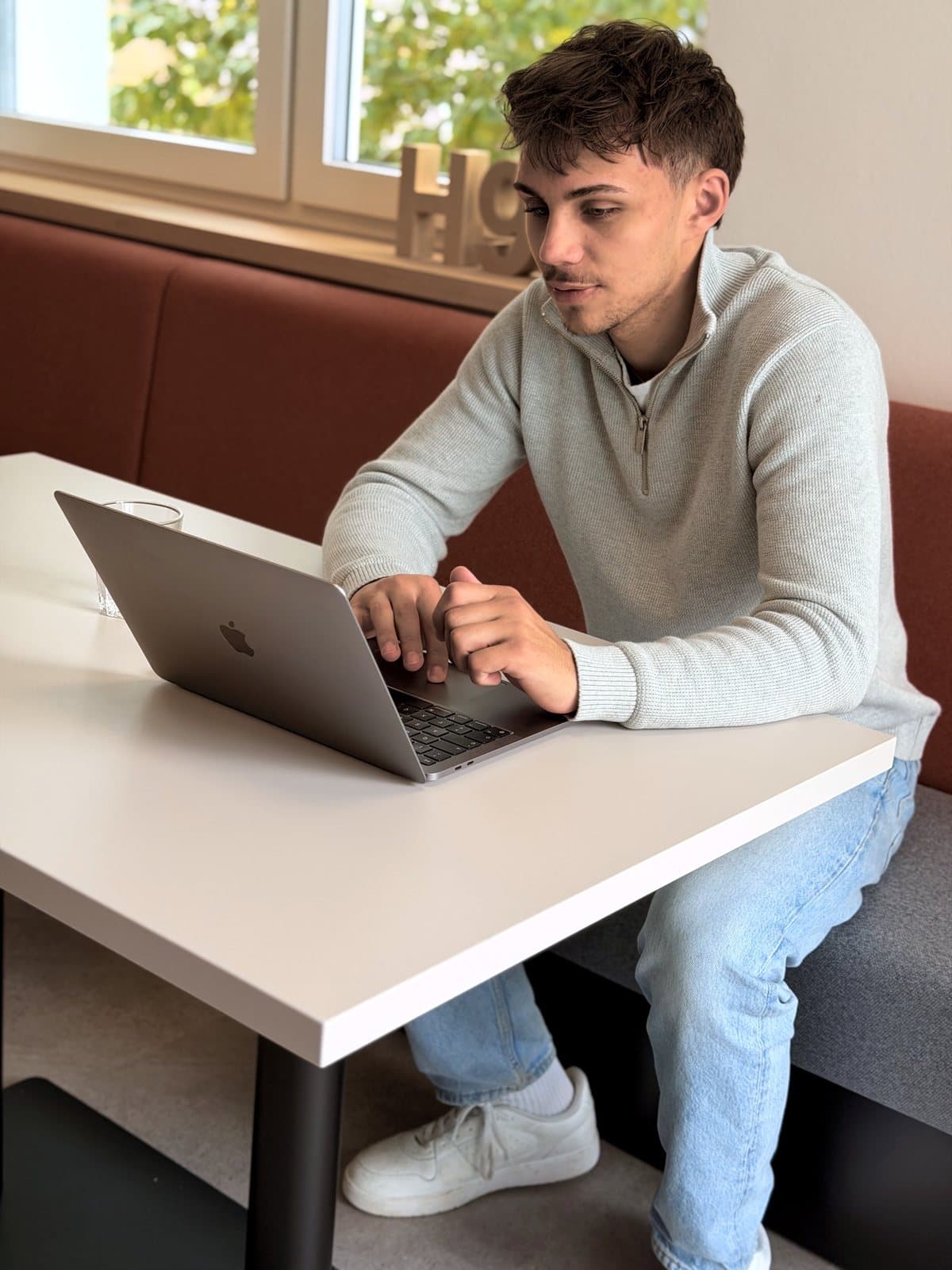 Luis Sabisch sitzt an einem hellen Tisch vor dem Laptop, daneben ein Glas Wasser.